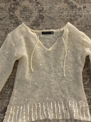 Vit stickad tröja med paljetter - Säljer en vit stickad tröja från Twilfit med v-ringning och knytband vid halsen. Tröjan har långa ärmar och är dekorerad med små vita paljetter över hela samt större paljetter längs nederkanten för extra bling. Finns ett mindre hål som syns på sista bilden! 