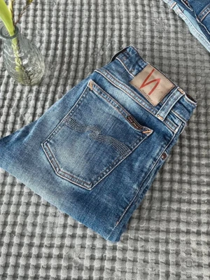 Blå slim fit jeans från Nudie Jeans - Snygga blå slim fit jeans från Nudie Jeans med klassisk femficksmodell och slitna detaljer. Jeansen har en ljus tvätt, kontrastsömmar och Nudie-logga på bakfickan. Tillverkade i bomull för en bekväm känsla och perfekt passform.
