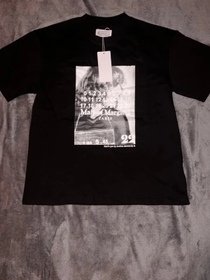 Maison Margiela  t-shirt - Svart t-shirt från Maison Margiela med stor grafisk print framtill i vitt och grått. Klassisk passform med rund hals och korta ärmar. Perfekt för dig som gillar streetstyle och vill sticka ut med en unik design.