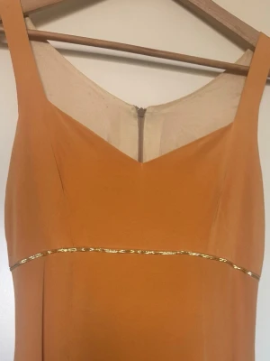 Skräddarsydd sidenklänning med gulddetalj - Elegant långklänning i persikofärg med v-ringad front och breda axelband. Klänningen har en dekorativ guldfärgad detalj under bysten och stängs med dragkedja i ryggen. Perfekt för balen. Skräddarsydd i finaste siden med fint foder. 
