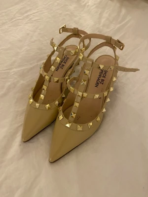 Beige pumps med nitar Shoe Biz - Snygga beige pumps från Shoe Biz Copenhagen med spetsig tå och dekorativa guldfärgade nitar på remmarna. Skorna har en T-rem design och justerbar ankelrem med spänne. Materialet är läder och de har en elegant och trendig look.