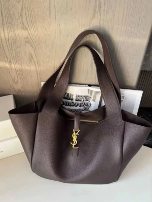 Mörkbrun YSL handväska i läder - Snygg mörkbrun handväska från Yves Saint Laurent i mjukt, strukturerat läder. Väskan har dubbla handtag och ett elegant guldigt YSL-emblem framtill. Perfekt storlek för att få plats med allt du behöver och ger en lyxig touch till din outfit.