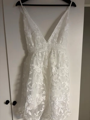 Vit spetsklänning med blommönster - Söt vit kortklänning med tunna axelband som korsas i ryggen och djup v-ringning både fram och bak. Klänningen har vackra spetsdetaljer och ett blommigt mönster i tyget. Midjan är markerad med en bred spetskant och kjolen är lätt utställd för en luftig känsla.