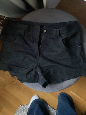 Svarta jeansshorts med rå kant - Säljer ett par svarta jeansshorts med rå, fransig kant och dragkedja framtill. Shortsen har dekorativa detaljer på bakfickorna. Perfekta för sommaren och ger en avslappnad vibe.