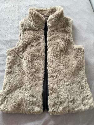 Beige fluffig pälsväst med dragkedja - Superskön beige pälsväst i fuskpäls med hög krage och dragkedja framtill. Västen har en mjuk och fluffig känsla och är perfekt för att addera lite extra stil till din outfit. Passar perfekt på våren och hösten när det är för varmt för jacka men för kallt för bara t-shirt!