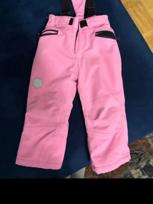 Rosa mjukisbyxor med hängslen - Säljer ett par rosa mjukisbyxor med svarta detaljer och justerbara hängslen. Byxorna har dragkedja fram, resår i midjan och reflexdetaljer på benet. Perfekta för chill dagar eller när du vill ha det bekvämt.