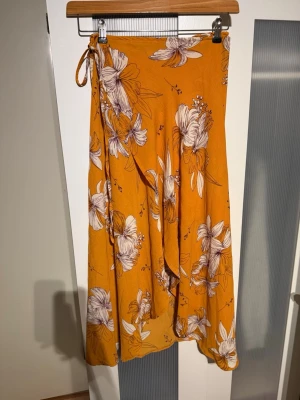 Blommig omlottkjol i orange - Säljer en snygg omlottkjol i orange med stora vita och lila blommor. Kjolen har knytband i midjan och ett flowigt fall som ger en avslappnad vibe. Perfekt för dig som gillar färg och mönster. Materialet känns lätt och passar bra till varmare dagar.
