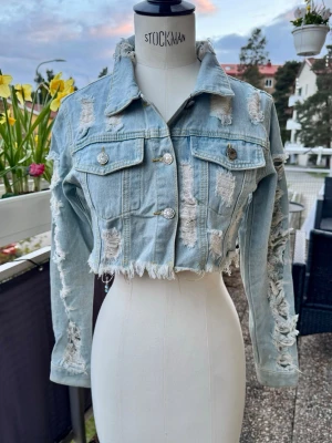Cropped ljusblå jeansjacka med slitningar - Cool cropped jeansjacka i ljusblå denim med råa kanter och kraftiga slitningar på både fram- och baksidan samt ärmarna. Jackan har två bröstfickor med knappar och klassisk krage. 