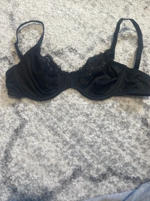 Svart spets-bh med tunna band - Aldrig använd, Säljer en svart bh med tunna axelband och vacker spetsdetalj över kuporna. Bh:n har en klassisk form med bygel och justerbara band. Materialet är en kombination av satin och spets som ger en elegant look.