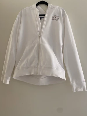 Vit hoodie med dragkedja från Tommy Hilfinger. - Snygg vit hoodie från Tommy Hilfiger med dragkedja framtill och två stora fickor. Klassisk logga på bröstet och ribbade muddar vid ärmslut och nederkant. Tillverkad i mjukt bomullsmaterial och har en huva för extra chill vibe.