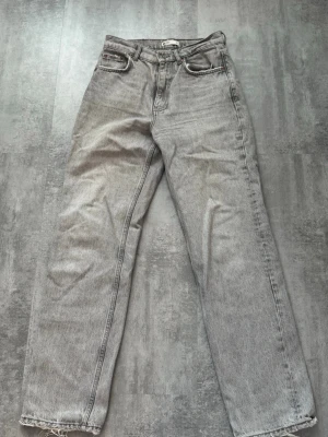 Grå raka jeans  - Säljer ett par grå jeans med rak passform och klassisk femficksdesign. Jeansen har hög midja och råa kanter längst ner på benen. Materialet är denim i bomull och färgen är ljusgrå med lätt tvättad look.
