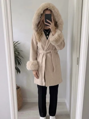 Beige kappa med pälsdetaljer  - Säljer en elegant beige kappa med äkta päls på huva och ärmslut. Kappan har en knytning i midjan och är perfekt för kyligare dagar. Den har en mjuk och lyxig känsla, vilket gör den till ett stilrent val för höst och vinter. S/M