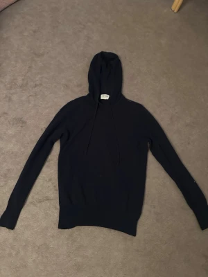 Svart hoodie i kashmir från Soft Goat - Mysig svart hoodie från Soft Goat i 100% kashmir. Klassisk modell med huva och dragsko, samt långa ärmar. Perfekt för dig som vill ha en stilren och lyxig hoodie med mjuk känsla. Tröjan är lite trasig precis vid halsen och har en pytteliten fläck längst ner men det ser man nästan inte. 💙