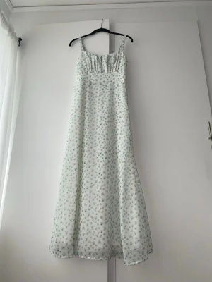 Lång blommig sommarklänning - Bustier Strap Dress, superfin långklänning med smala axelband och småblommigt mönster i ljusa färger. Klänningen har en lätt och luftig känsla, perfekt för varma dagar. Den är vit i grunden med gröna och blå blommor och har en söt rynkad detalj vid bysten.
