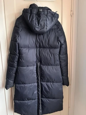 Svart lång dunjacka från uhip  - Säljer en svart lång dunjacka från uhip  med huva och dragkedja framtill. Jackan har två fickor med lock och logotyp på bröstet samt ärmen. Perfekt för kalla vinterdagar, håller dig varm och bekväm. Materialet är glansigt och quiltat.