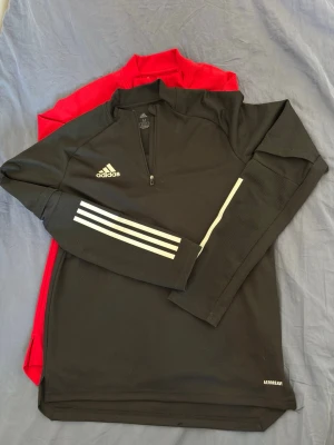 Adidas träningsöverdelar svart och röd - Två långärmade träningsöverdelar från Adidas, en svart med vita detaljer och dragkedja samt en röd med dragkedja. Båda har Adidas-loggan på bröstet och är tillverkade i ett lätt och funktionellt material som passar perfekt för sport och träning.