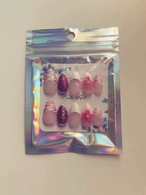 Press-on naglar rosa och vinröd - Handgjorda press-on naglar i mandelform med mix av rosa, vinröda och vita nyanser. Några naglar har dekorativa ränder, andra har blommönster och silverdetaljer. Perfekt för dig som vill ha unika och trendiga naglar utan att gå till salong.