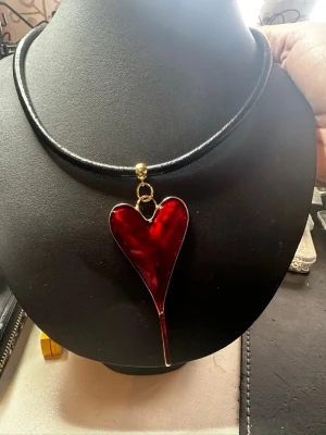 Halsband med rött hjärthänge - Unikt halsband med svart läderrem och ett stort, blankt rött hjärthänge i metall med guldfärgade detaljer. Hjärtat har en modern, utdragen form och hänger i en guldfärgad ring. Perfekt för dig som vill sticka ut med ett statement-smycke.