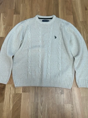 Beige kabelstickad tröja U.S. Polo Assn. - Kabelstickad beige tröja från U.S. Polo Assn. med rund halsringning och broderad logga på bröstet. Tröjan har ribbade muddar vid ärmslut och nederkant. Perfekt för dig som gillar klassisk och stilren design. Passar som m/L