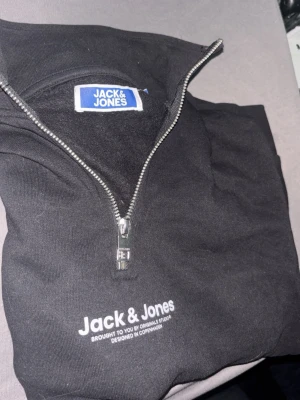 Svart halfzip hoodie från Jack & Jones - Snygg svart hoodie från Jack & Jones med halv dragkedja framtill och vit logga på bröstet. Tillverkad i mjukt bomullsmaterial och har en klassisk passform. Perfekt för en chill och avslappnad stil.
