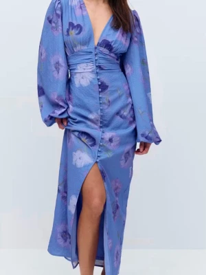 Blommig blå långklänning med slits - Långklänning i blått med lila och vita blommor, v-ringad och med markerad midja. Klänningen har långa puffärmar, knappar framtill och en hög slits. Perfekt för dig som vill sticka ut med en romantisk look.