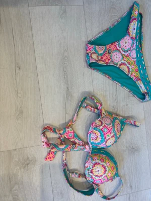 Färgglad blommig bikini med knyt - Säljer en bikini med färgglatt blommigt mönster i rosa, gul, grön och turkos. Överdelen har vadderade kupor och knyt i nacken samt ryggen. Underdelen har samma mönster. Märket är oklart, köpt på en marknad i Frankrike!