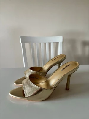 Guldiga klackar från Boohoo - Säljer ett par guldiga klackar från Boohoo med öppen tå och smal hög klack. Skorna har en bred rem över foten och är tillverkade i ett glansigt material som ger en lyxig känsla. Perfekta för dig som vill sticka ut med metallic på fötterna.