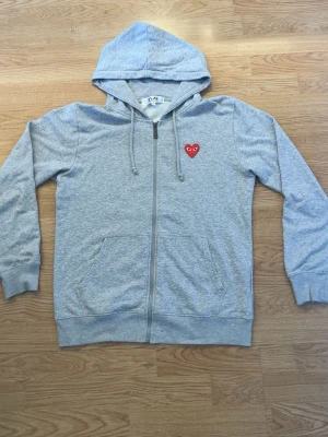 Grå hoodie från Play Comme des Garçons - Snygg grå zip-hoodie från Play Comme des Garçons med det klassiska röda hjärtat på bröstet. Hoodien har dragkedja, två fickor fram och justerbar huva med snören. Tillverkad i mjuk bomullsblandning och har ribbade muddar vid ärmar och nederkant. Strl L men passar även M också 
