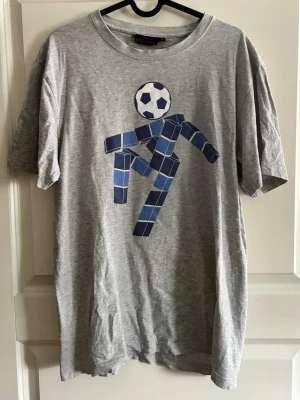 Grå t-shirt med blått fotbollstryck - Säljer en grå t-shirt från järnkaminerna med ett unikt tryck av en figur som spelar fotboll i blå toner på framsidan. T-shirten har rund hals och korta ärmar. Perfekt för dig som gillar sportiga och kreativa motiv.