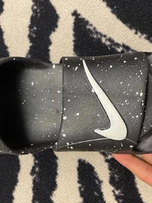 Nike Vapormax svarta tofflor - Coola svarta Nike Vapormax tofflor med vit swoosh och vita stänk över hela ytan. Tofflorna har en unik, chunky sula och bred rem över foten. Perfekt för dig som gillar streetstyle och vill ha något bekvämt och trendigt. Materialet är syntetiskt och designen är riktigt modern.