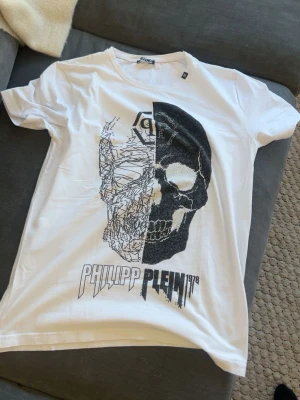Vit PP t-shirt med döskalle - Snygg vit t-shirt med ett stort tryck av en döskalle framtill i svart och vitt. T-shirten har korta ärmar, rund halsringning. Märkesdetaljer syns både fram och bak. Perfekt för dig som gillar streetwear och statement-plagg. Står xl på men den passar M och L