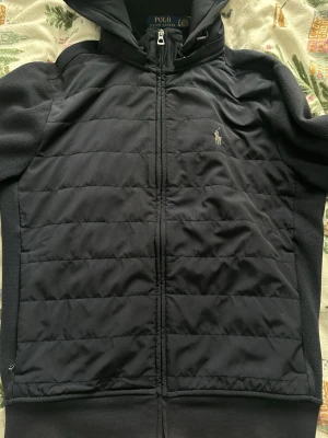 Svart hoodie från Polo Ralph Lauren - Svart hoodie med quiltad framsida och dragkedja från Polo Ralph Lauren. Klassisk huva och broderad logotyp på bröstet. Materialmix med slät och quiltad yta, perfekt för en cool och avslappnad look.