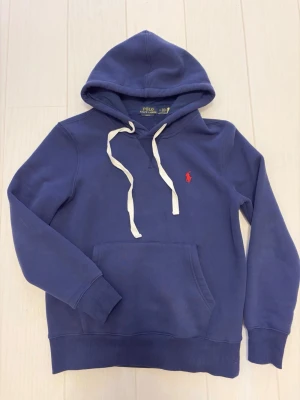 Blå hoodie från Polo Ralph Lauren - Mörkblå hoodie från Polo Ralph Lauren med vita band och röd logga på bröstet. Hoodien har känguruficka framtill, ribbade muddar och är tillverkad i mjuk bomullsblandning. Denna är i storlek S och är använd ett fåtal gånger. Längden på plagget är ungefär 61cm. Jättefin navy färg som passar till mycket. 