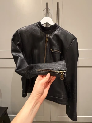 Svart skinnjacka med dragkedjor - Snygg svart skinnjacka från Jofama i äkta läder. Jackan har ståkrage med tryckknapp, dragkedja framtill, bröstficka med dragkedja och dragkedjor vid ärmsluten. Perfekt för en cool och stilren look.