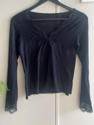 Svart långärmad topp med spets - Marinblå långärmad topp med v-ringning och spetsdetaljer både vid halsringningen och ärmsluten. Tunn och mjuk i materialet, perfekt för att styla lager på lager eller bära som den är. Enkel men ändå elegant design.