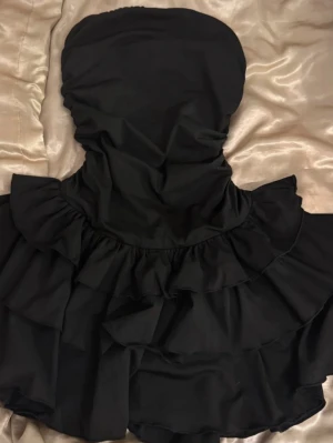 Svart bandeau klänning med volanger - Säljer en snygg svart bandeau klänning med volanger. Axelbandslös modell som ger en trendig look.💕 ingen lapp men stod xs vid köp och sitter som xs. Köpt i Spanien så osäker på märke. Skickar snabbt💌