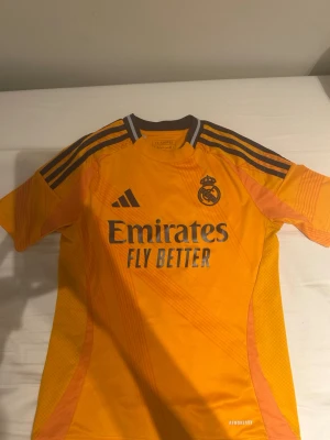 Real Madrid Valverde matchtröja - Säljer en gul-orange Real Madrid matchtröja från Adidas med Valverde och nummer 8 på ryggen. Tröjan har grå detaljer, tre ränder på axlarna och sponsorlogga på bröstet. Materialet är lätt och andas, perfekt för fotboll och träning.