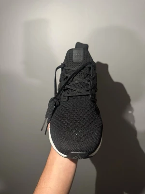 Svarta Adidas Ultra Boost sneakers - Säljer ett par svarta Adidas Ultra Boost sneakers med stickad ovandel och vita Boost-sulor. Skorna har klassisk låg profil, svarta snören och diskret Adidas-logga på plösen. Perfekta för dig som gillar stilrena och bekväma sneakers med sportig känsla.