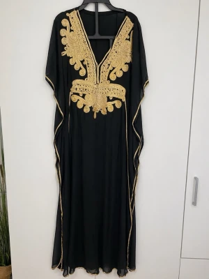 Svart långklänning med guld broderi - Elegant svart kaftan/klänning med vackra gulddetaljer, direkt från Marocko. Rikligt broderad framtill med dekorativa mönster som ger en lyxig och autentisk look. Lös och bekväm passform som passar perfekt för fest, högtider eller semestrar.  Storlek: passar XXS–S.  Lätt och luftig – både stilren och bekväm att bära.  Fint skick.