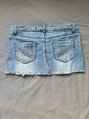 Blå jeanskjol - Säljer en blå jeanskjol med slitna detaljer, rå kant och dekorativa sömmar på bakfickorna. Kjolen har fem fickor, bälteshällor och knappstängning framtill. Perfekt för dig som gillar en edgy och avslappnad stil.