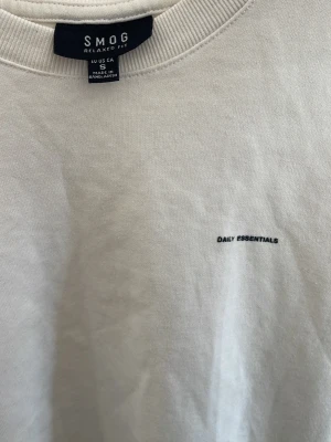 Vit t-shirt från SMOG Relaxed Fit - Vit t-shirt från SMOG Relaxed Fit med minimalistisk text 'DAILY ESSENTIALS' på bröstet. Klassisk rund halsringning och korta ärmar. Tillverkad i mjuk bomull för en skön och avslappnad känsla.