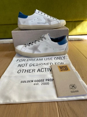 Golden Goose vita sneakers med blå detalj - Golden Goose sneakers i vitt läder med grå stjärndetalj på sidan och blå hälkappa. Klassisk låg modell med grå skosnören och beige sula. Snygg och stilren design som passar till många outfits. Storlek 41 men passar 42. Kommer med originalpåse och pass.