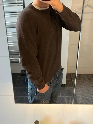Mörkbrun rundhalsad tröja - En enkel och stilren mörkbrun tröja med rund halsringning och långa ärmar. Tröjan har ribbade muddar vid ärmslut och nederkant, perfekt för lager på lager och passar till jeans. Materialet ser mjukt och bekvämt ut.