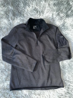 Mörkgrå stickad tröja Arc'teryx - Snygg mörkgrå stickad tröja från Arc'teryx med halv dragkedja framtill och hög krage. Tröjan har lång ärm och en diskret logga på ärmen. Perfekt för lager-på-lager och outdoor-vibe. Materialet är mjukt och stretchigt för extra komfort.