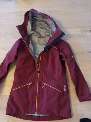 Vinröd vindjacka från Peak Performance  - Snygg vinröd vindjacka med längre passform och huva från Hällräven. Jackan har orange dragkedjor, flera praktiska fickor med dragkedja och diskret logotyp på bröstet. Materialet är slitstarkt och vattenavvisande, perfekt för uteliv och äventyr.