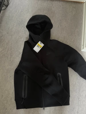 Svart Nike hoodie med dragkedja - Säljer en svart hoodie från Nike med hel dragkedja framtill och två fickor med dragkedja. Hoodien har en stilren design med diskret Nike-logga på bröstet och justerbar huva. Tillverkad i mjukt och bekvämt material, perfekt för en avslappnad look.