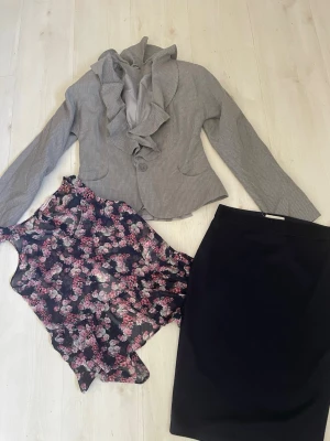 Grå kavaj med volangkrage+Kjol +Linne  - Säljer en grå kavaj med diskret randigt mönster och snygg volangkrage. Kavajen har knappar framtill och figurnära passform. Perfekt att matcha med en blommig topp och svart kjol för en stilren look.  Kjol i blått i bomull  och Tunnt Linne i blommigt 