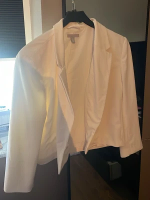 Vit kavaj från H&M - Stilren vit kavaj från H&M med klassisk krage och öppen front. Kavajen har en rak passform och är tillverkad i ett mjukt tyg som ger en snygg och modern look. Perfekt att matcha med jeans eller kjol för en trendig outfit.