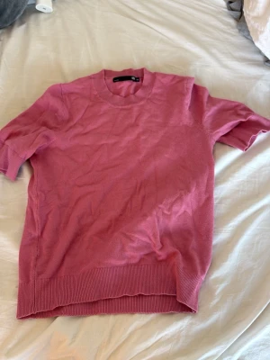 Rosa stickad t-shirt från lager 157 - Säljer en rosa stickad t-shirt från lager 157 med korta ärmar och rund halsringning. Tröjan har ribbade muddar vid ärmslut och nederkant, vilket ger en snygg och avslappnad look. Perfekt för dig som gillar stilrena och bekväma plagg.