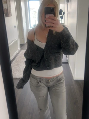 Grå cropped kofta med knappar - Säljer en grå, cropped kofta med v-ringning och knappar framtill. Koftan har två dekorativa fickor med knappdetaljer och är stickad i ett mjukt material. Perfekt för lager på lager och en avslappnad look.